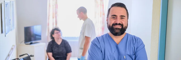 Portraitaufnahme einer männlichen Pflegekraft. Im Hintergrund eine Pflegeszene mit einer Patientin
