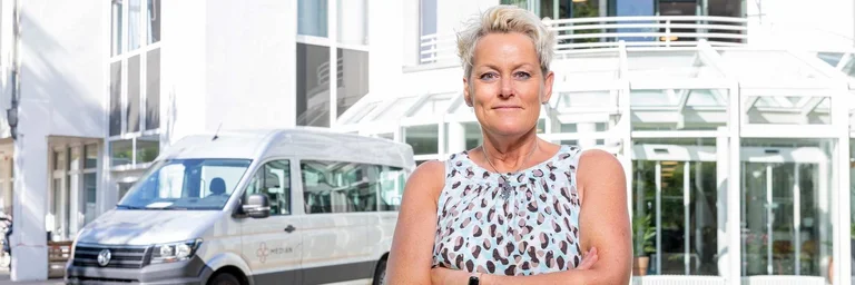 Eine Mitarbeiterin des Fahrdienst steht vor einem PKW und dem Klinikgebäude