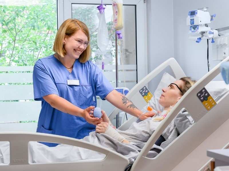 Eine Mitarbeterin ist bei einer Patientin im Pflegebett und machen Greifübungen mit einem Igelball