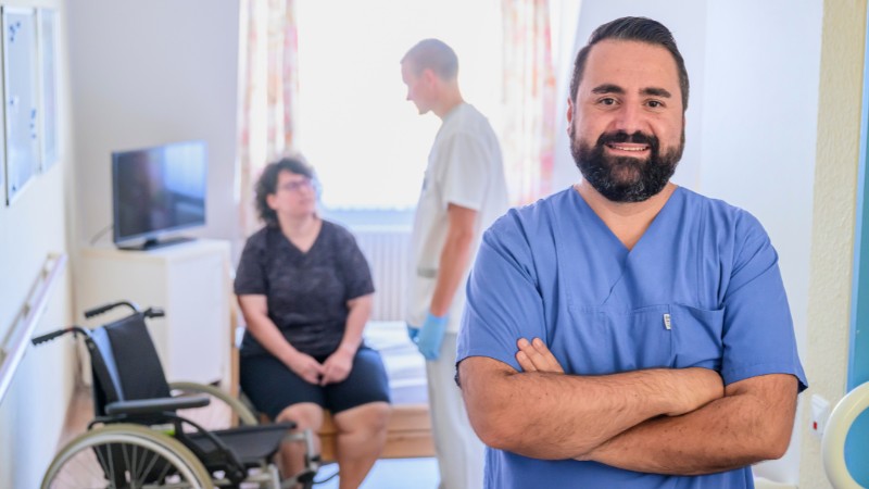 Portraitaufnahme einer männlichen Pflegekraft. Im Hintergrund eine Pflegeszene mit einer Patientin
