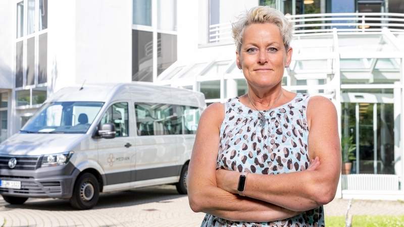 Eine Mitarbeiterin des Fahrdienst steht vor einem PKW und dem Klinikgebäude